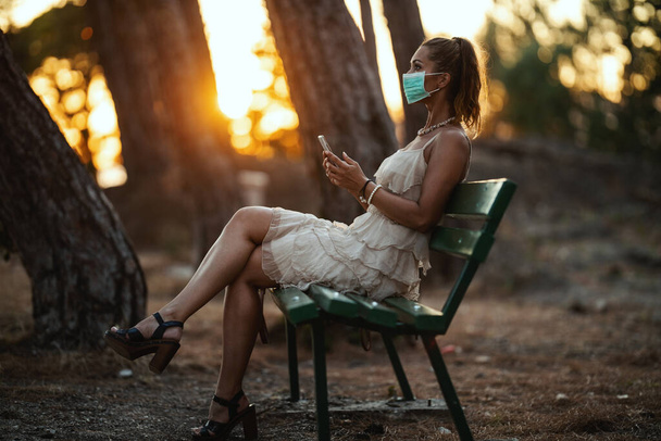 Aufnahme einer attraktiven jungen Frau, die eine chirurgische Maske trägt und ein Smartphone benutzt, während sie sich im Park isoliert. - Foto, Bild