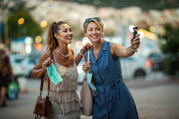 Girato di due attraenti amiche donne che indossano una maschera chirurgica e utilizzano una fotocamera digitale mentre esplorano la meravigliosa città di Meditteranian. - Foto, immagini