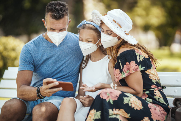 Self-isolation in nature. Young happy family with protective N95 mask having fun during walk in the park and making video call with smartphone. - Photo, Image