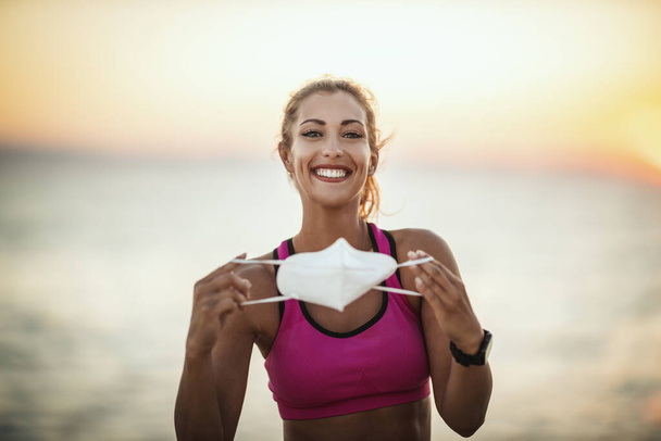 Retrato de una corredora sonriente con máscara protectora N95 mientras entrenaba cerca de la playa al atardecer durante la pandemia de COVID-19. - Foto, Imagen