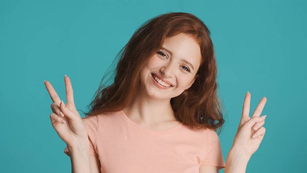 Pretty smiling redhead woman joyfully showing peace gesture on camera over colorful background. Victory sign - Photo, Image