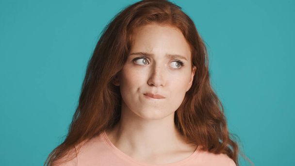 Beautiful serious redhead girl biting lips thoughtfully looking away over colorful background. Nervous  expression - 写真・画像