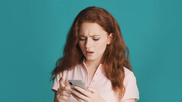 Atrakcyjny zdenerwowany ruda dziewczyna frustrująco za pomocą smartfona na kolorowe tło. Smutne wyrażenie - Zdjęcie, obraz