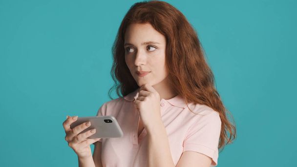 Atrakcyjny marzycielski ruda dziewczyna starannie oglądanie wideo na smartfonie na kolorowe tło. Wyrażenie myślenia - Zdjęcie, obraz