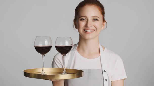Atraktivní usmívající se servírka v zástěře podnos s vínem šťastně dívá do kamery přes bílé pozadí - Fotografie, Obrázek