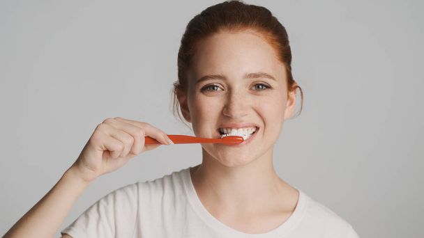 Güzel kızıl saçlı kızın portresi dikkatle kameraya bakıyor beyaz arkaplanda dişlerini fırçalıyor - Fotoğraf, Görsel