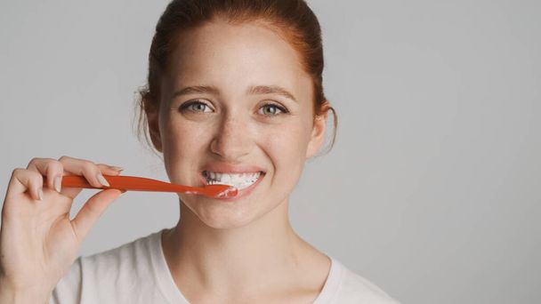 Portret van mooie roodharige meisje gelukkig poetsen tanden kijken in camera over witte achtergrond - Foto, afbeelding