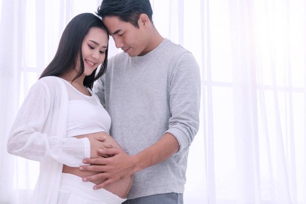 Ehemann umarmt seine Frau und formt mit ihren Händen Herz auf dem Schwangerschaftsbauch. Glückliches Paar junger Mann umarmt seine schwangere schöne Frau, lächelt und schaut sich zu Hause in die Augen. - Foto, Bild