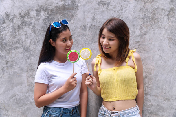 Lifestyle concept. Happy smile two young women holding lollipop looking each other on grey background. Cheerful girl with sunglasses holding candy in her hand looking at her friend. - Photo, Image