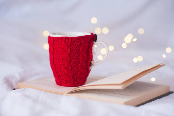 Knitted cup of tea staying on open book over Christmas lights at background close up. Winter holiday season. Xmas.  - Photo, Image