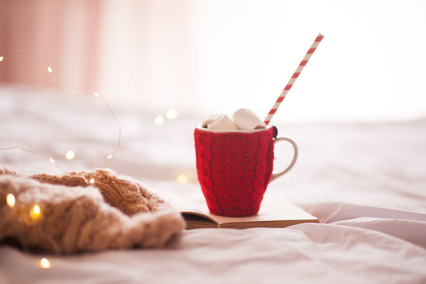 Šálek kávy s červenou pletenou textilií s marshmallow a vodní dýmkou v posteli zblízka. Zimní prázdniny. Vánoce. Dobré ráno.  - Fotografie, Obrázek