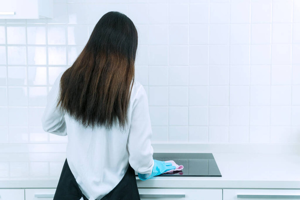 La espalda de una mujer joven con guantes protectores azul limpiando el polvo con aerosol y tela mientras limpia su cocina. Concepto de personas, tareas domésticas y limpieza. - Foto, imagen