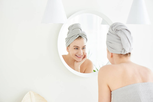 Portrait of  young girl with  towel on head in white bathroom looks and touches her face in the mirror and enjoys youth and hydration. Natural beauty, home care for problem skin - Valokuva, kuva