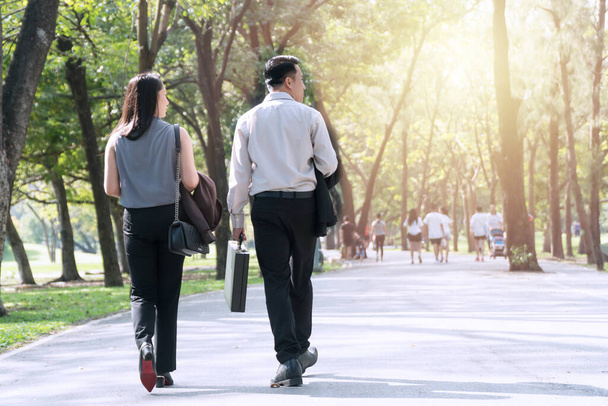 The back of couple businessman and businesswoman walking together within the park relaxed after work. Two businessmen holding cup of coffee walk talk happily in the park. Relaxing concept - Photo, Image