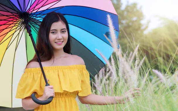 Hymyilevä nuori nainen pitämällä sateenkaaren sateenvarjo kukka ruoho kenttä katselee kameraa luonnollisella taustalla. Matkustaa rentoutua aika käsite. - Valokuva, kuva