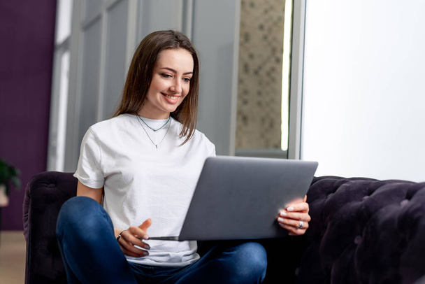 Young beautiful woman using a laptop computer at home. Black sofa. Smiling young woman with laptop. - Fotó, kép