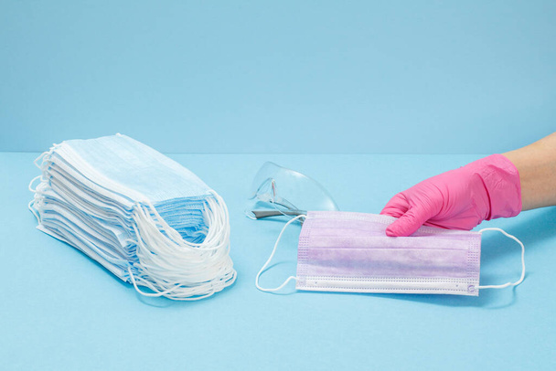 Vrouwelijke arts in een latex handschoen met medische beschermende maskers in zijn hand en een stapel maskers, bril op een blauwe rug. Persoonlijke beschermingsmiddelen tegen virale infecties. - Foto, afbeelding