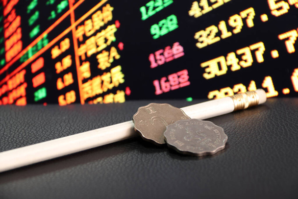 Две монеты по два гонконгских доллара с белым карандашом на черном полу и цифровая доска обмена валюты на заднем плане, концепция финансов. - Фото, изображение