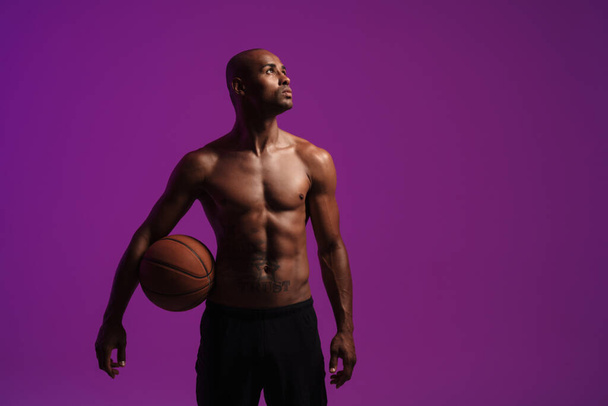 Photo of a serious strong african fitness sports man posing isolated over purple wall background while holding basketball - 写真・画像