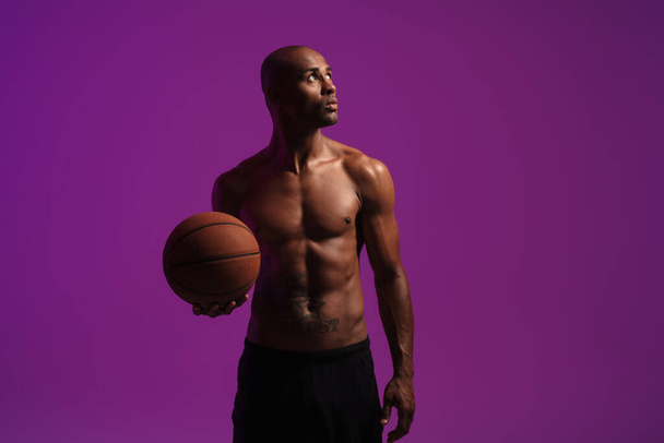 Picture of a concentrated strong african fitness sports man posing isolated over purple wall background while holding basketball - Photo, Image