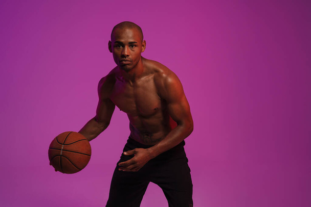 Image of a concentrated strong african fitness sports man isolated over pink wall background playing basketball - Фото, изображение