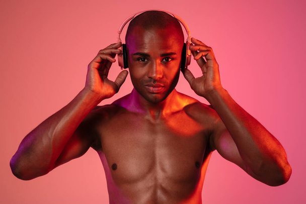 Image of african fitness man posing isolated over neon wall background while listening music with headphones - Zdjęcie, obraz
