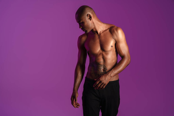 Image of athletic half-naked african american man with tattoo posing on camera isolated over purple wall - Fotó, kép
