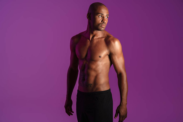 Image of athletic half-naked african american man with tattoo posing on camera isolated over purple wall - Foto, Imagem