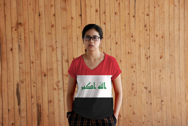 Жінка з іракським прапором колір сорочки і стояти з двома руками в пишних кишенях на дерев'яному фоні стіни, червоний білий і чорний з Богом найбільший в зеленому.  - Фото, зображення