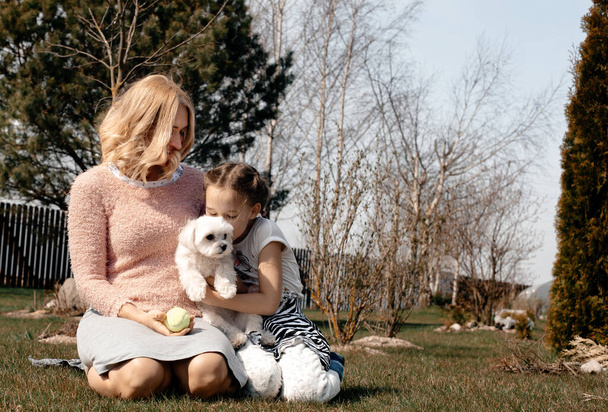 A little girl and her mother are sitting on the travet, playing with a small white dog, against the backdrop of the country garden. Carefree childhood. Pet care concept. Summer sunny day. - Foto, afbeelding