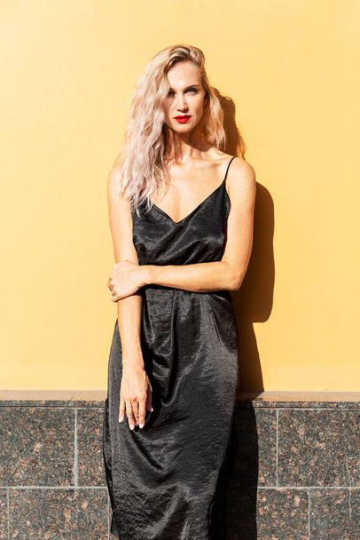 A young woman stands against a yellow wall of a building. Beautiful blonde in a black dress in bright sunlight. Vertical. - Foto, Bild