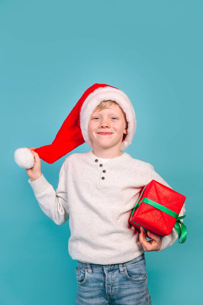 funny child in Santa red hat holding Christmas gift in hand on blue background. Christmas concept. - Foto, Imagen