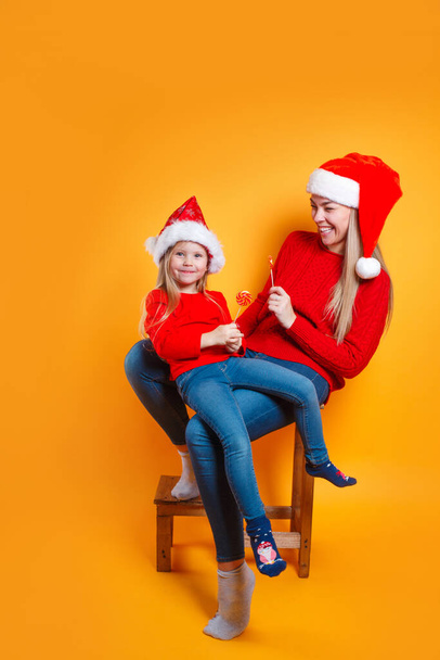 Cheerful young mother and cute daughter girl in Santa hats holding Chistmas candy on yellow background - Fotó, kép