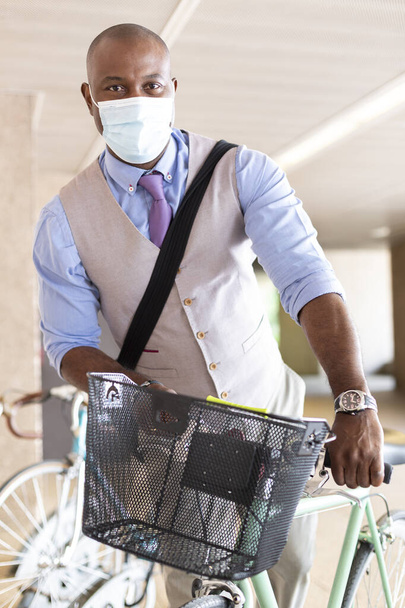 Schwarzer Unternehmer parkt sein Fahrrad. Er trägt eine medizinische Maske und formelle Kleidung. Konzept von gesundem Leben und neuer Normalität. - Foto, Bild