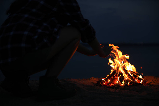 Woman warming hands near burning firewood on beach at night, closeup - Foto, imagen