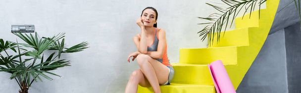 Panoramic orientation of sportswoman sitting on stairs near fitness mat at home - Fotografie, Obrázek
