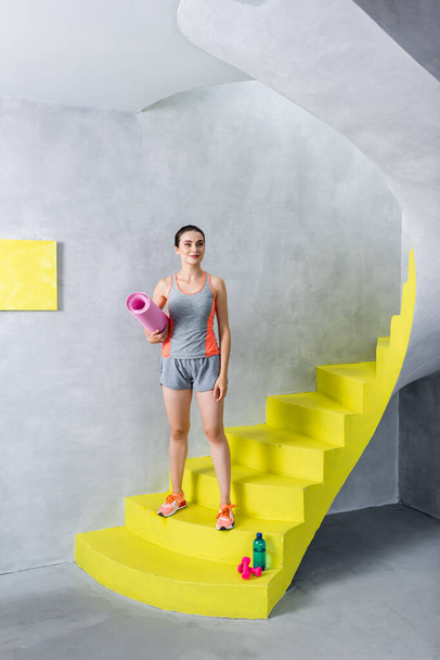 Młoda kobieta w odzieży sportowej trzyma matę fitness w pobliżu butelki wody i hantli na schodach w domu  - Zdjęcie, obraz