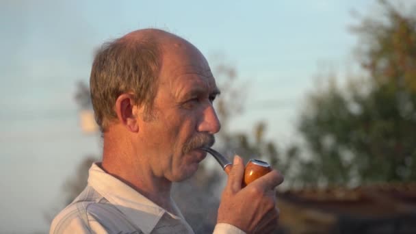 Vidéo au ralenti de Tabagisme dans une pipe à fumée épaisse.Dépendance à la nicotine chez les hommes d'âge moyen Un homme blanc avec une moustache allume une pipe en gros plan le soir au coucher du soleil à l'heure dorée - Séquence, vidéo