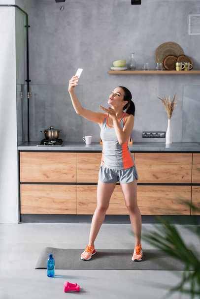 Selective focus of young sportswoman blowing air kiss near water and sport equipment in kitchen  - Foto, Bild