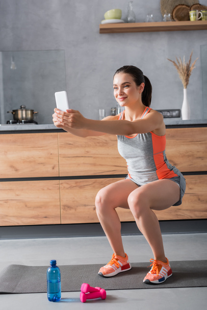 Selective focus of sportswoman doing squat while holding smartphone near water and dumbbells in kitchen  - Foto, imagen
