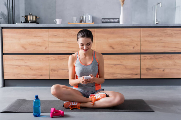 Sportswoman using smartphone on fitness mat near bottle of water and dumbbells in kitchen  - Фото, изображение