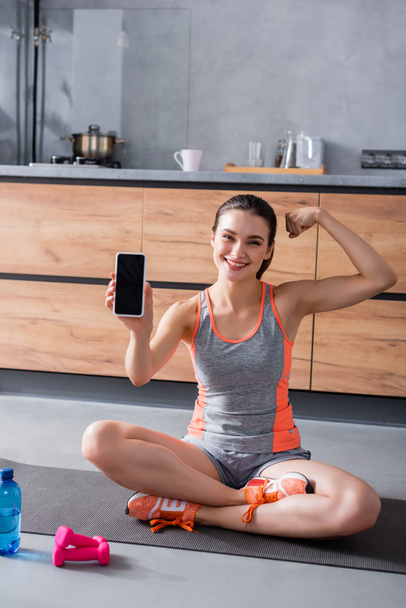 Sportswoman showing smartphone with blank screen near bottle of water and sport equipment in kitchen  - Zdjęcie, obraz