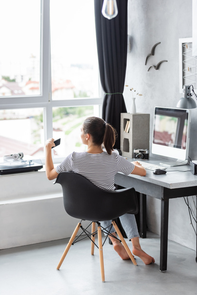 Takaisin näkymä nuori nainen käyttää älypuhelinta tietokoneen lähellä pöydällä kotona  - Valokuva, kuva