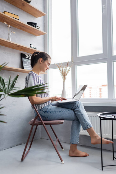 Side view of barefoot freelancer using laptop near window at home   - Foto, Imagem