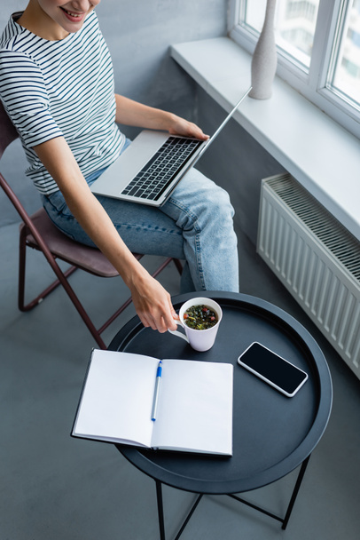 Vágott kilátás szabadúszó gazdaság csésze kávé használata közben laptop közelében okostelefon és notebook otthon  - Fotó, kép