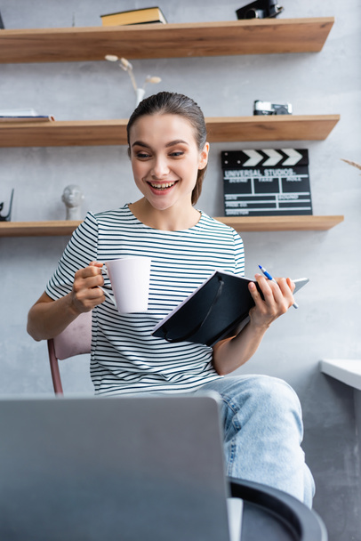 Selective focus of brunette teleworker with cup and notebook looking at laptop while working from home  - Φωτογραφία, εικόνα
