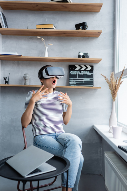 Selective focus of shocked woman in vr headset sitting near laptop on coffee table in living room  - Photo, Image