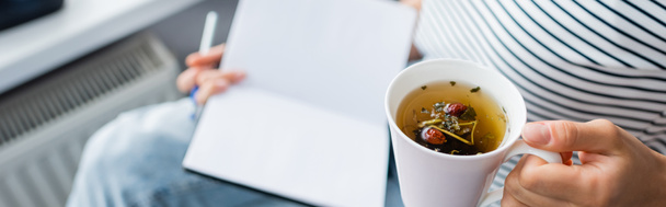 Panoramic shot of woman holding cup of herbal tea and notebook at home  - Foto, afbeelding