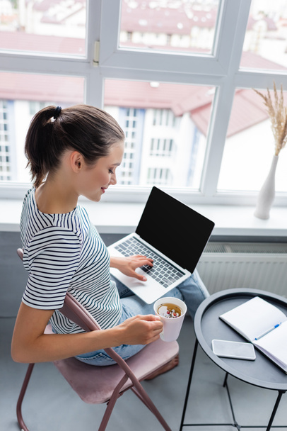 Bruneta na volné noze pomocí notebooku, zatímco drží šálek bylinného čaje v blízkosti notebooku a smartphone na stole   - Fotografie, Obrázek