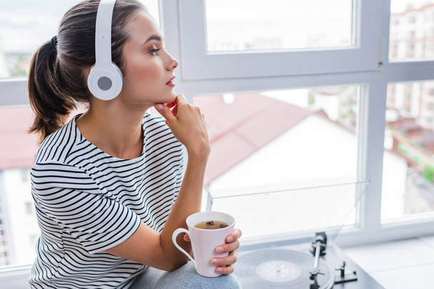 Young woman with cup of tea listening music in headphones near vinyl player on windowsill  - Photo, Image